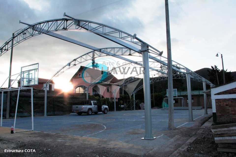Cancha exterior con estructura de acero al atardecer, con una camioneta blanca estacionada y edificios cercanos.