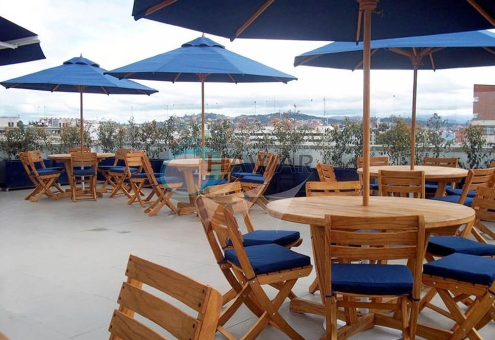Cafetería al aire libre en la azotea con mesas de madera, sombrillas azules y vistas a la ciudad al fondo.