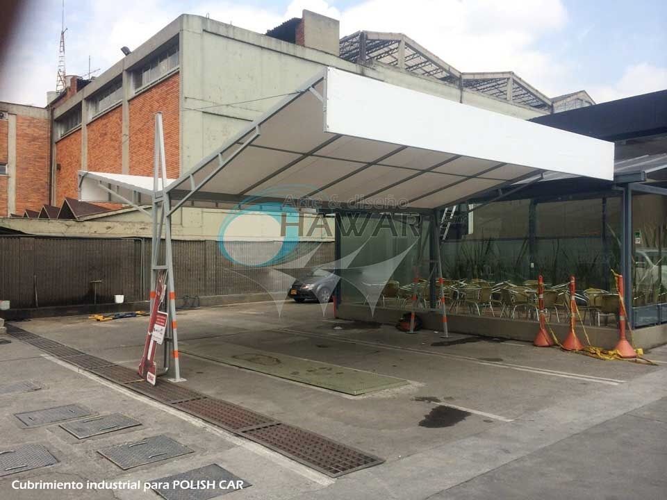 Zona de aparcamiento cubierta junto a un edificio industrial, con conos y un coche bajo una marquesina blanca.