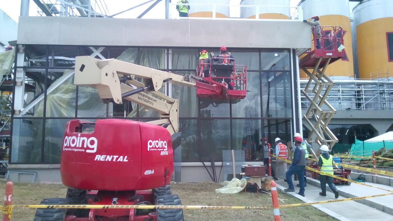 Un obrero de la construcción, con casco amarillo y chaleco de seguridad naranja, instala un gran panel de vidrio en un edificio de gran altura.