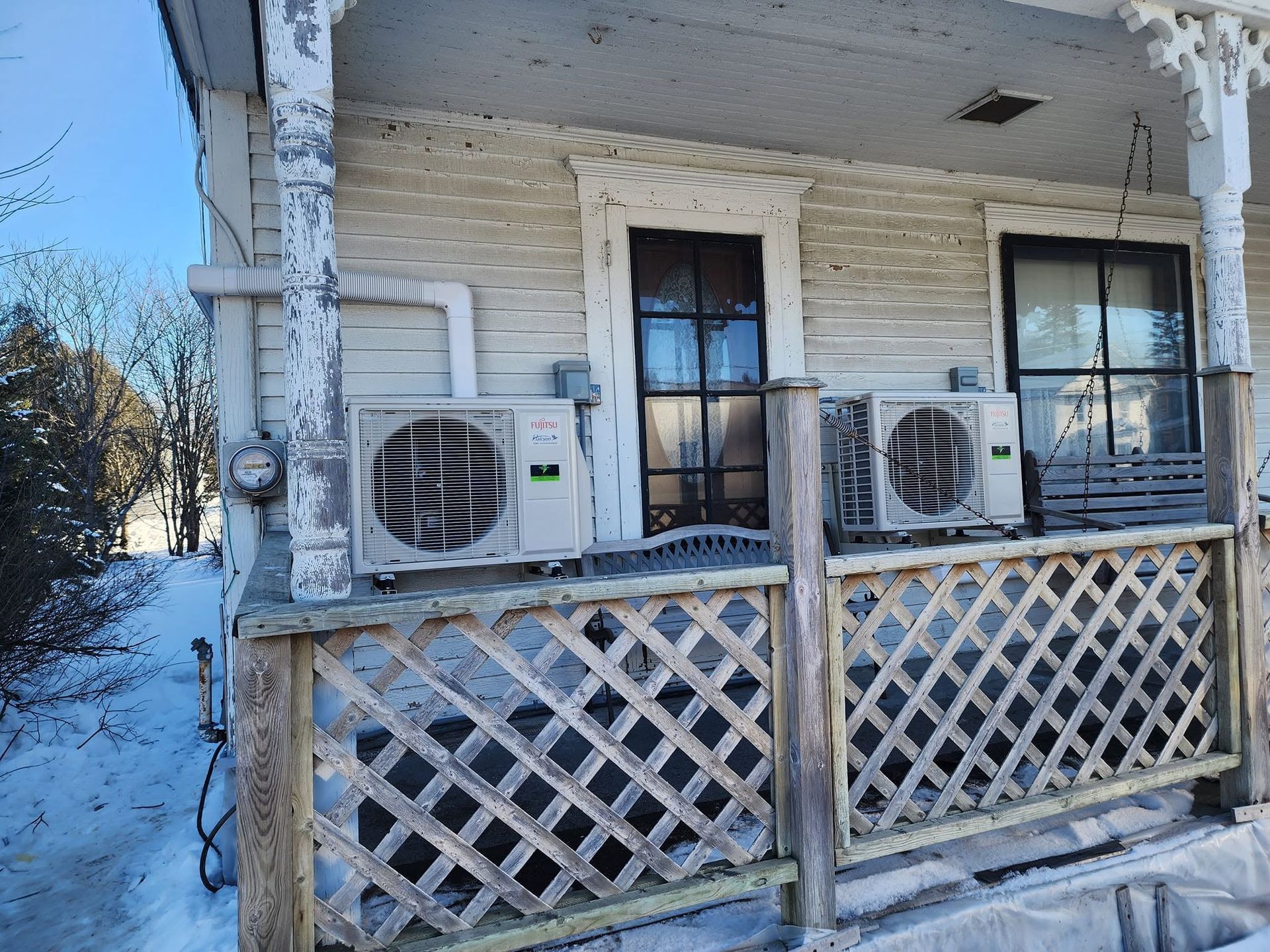 Snow-covered house porch with two wall-mounted air conditioners behind a lattice railing