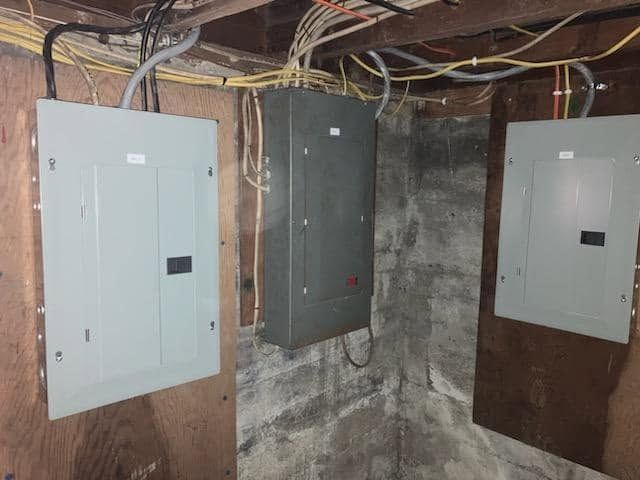 Basement wall with three electrical breaker panels and exposed wiring on concrete walls