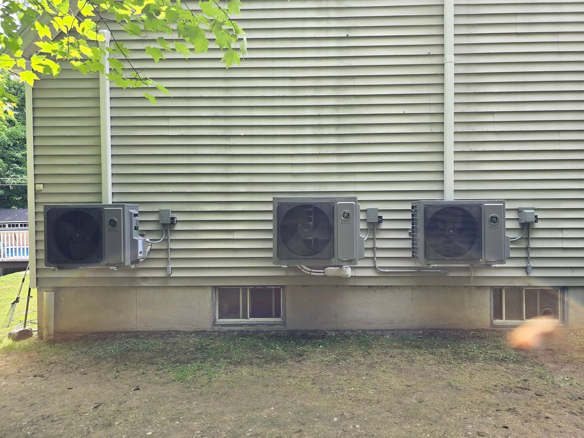 Side of a house with four exterior air conditioning units mounted along the wall under green leaves.