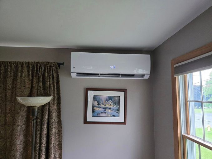 Living room wall with white air conditioner above a framed painting, beside a lamp and window.