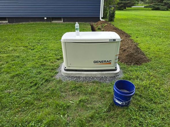 Outdoor generator on a concrete pad beside a blue bucket on a grassy lawn near a house