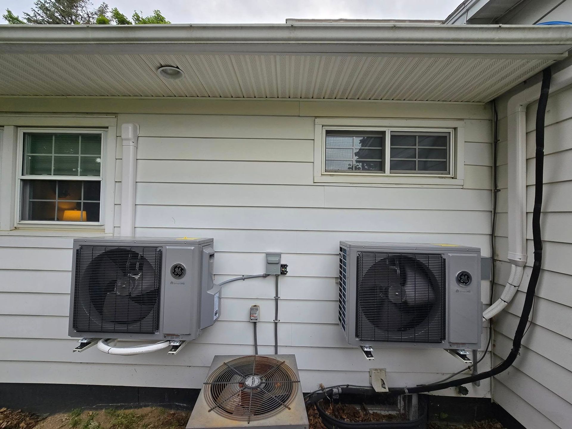 Two gray outdoor HVAC units mounted on a house exterior under a window and eaves