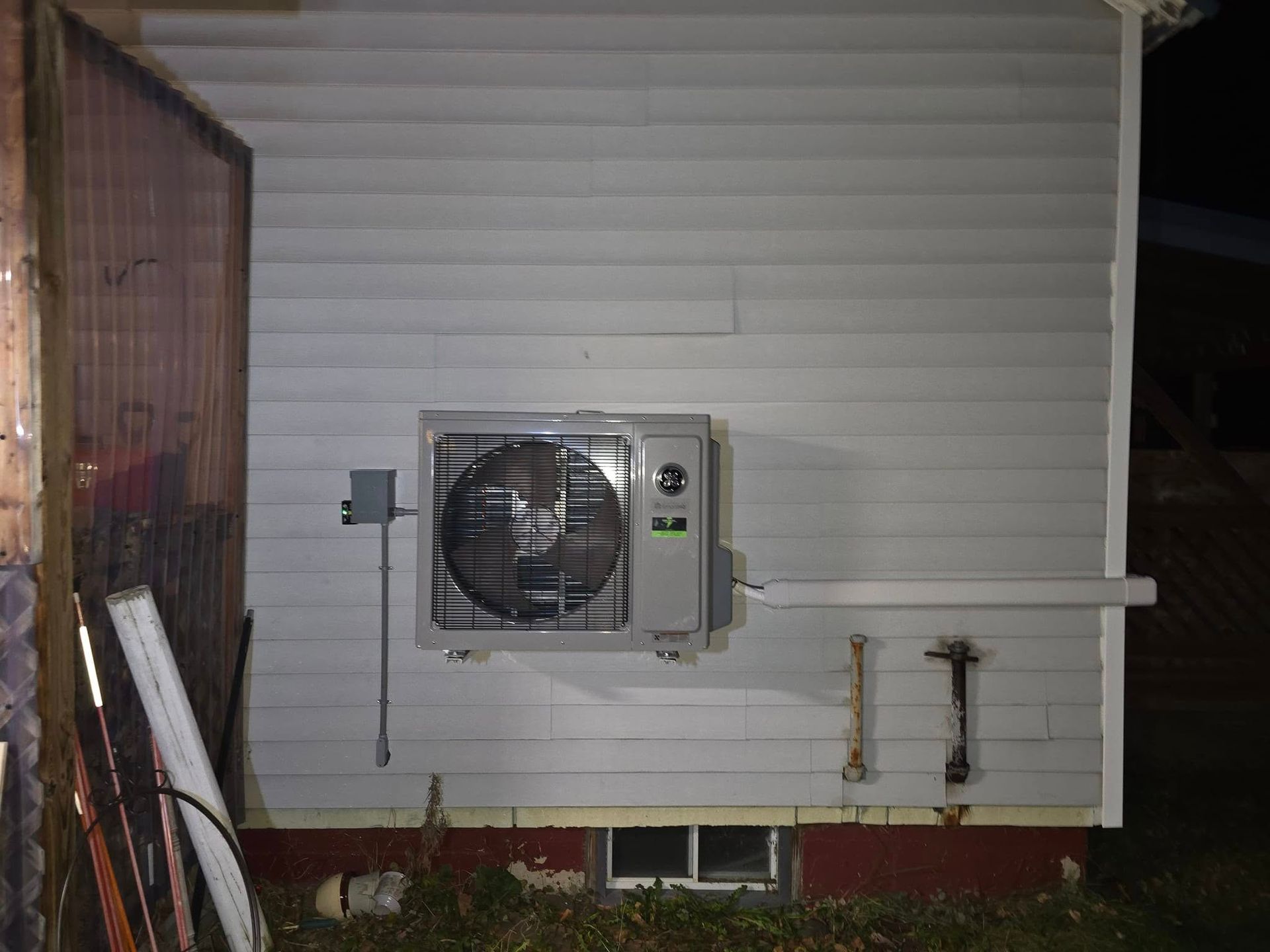Wall-mounted air conditioner on white siding beside a metal gate and pipes at night