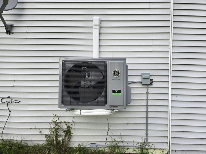 Outdoor air conditioning unit mounted on a house’s beige vinyl siding, with visible piping and electrical box