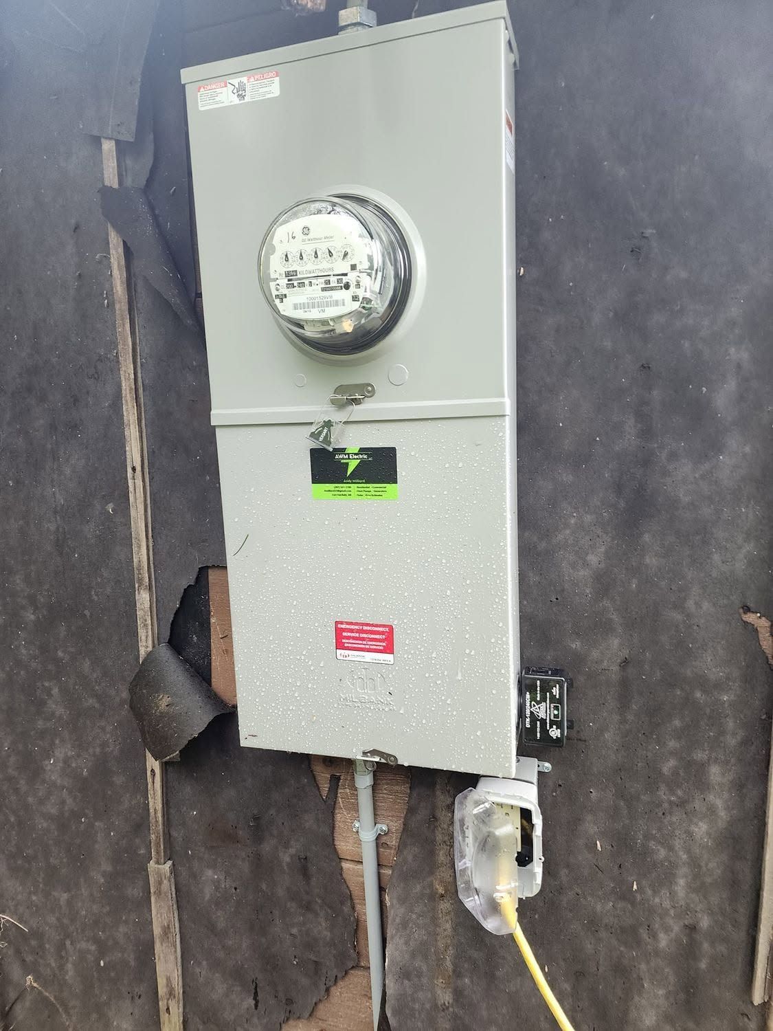 Gray electrical meter box mounted on a wall with conduit and outlet below
