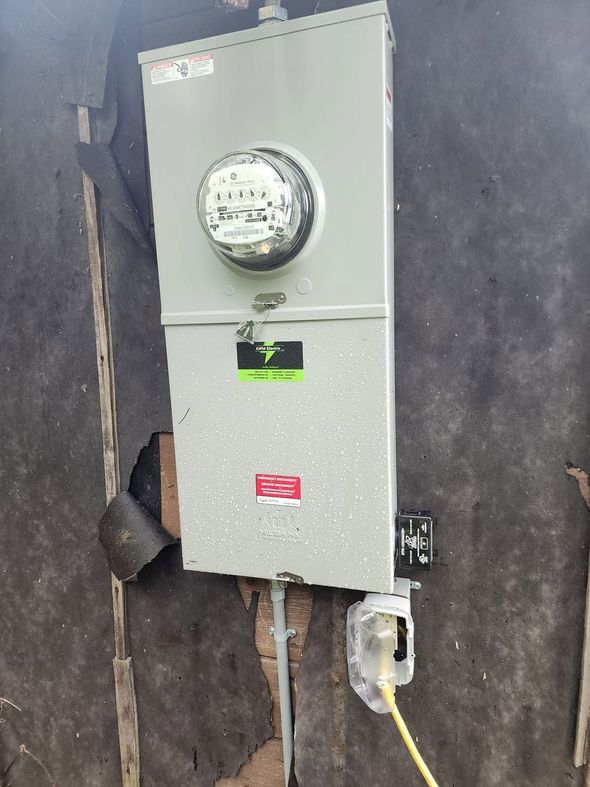 Gray electrical meter box mounted on a wall with conduit and outlet below