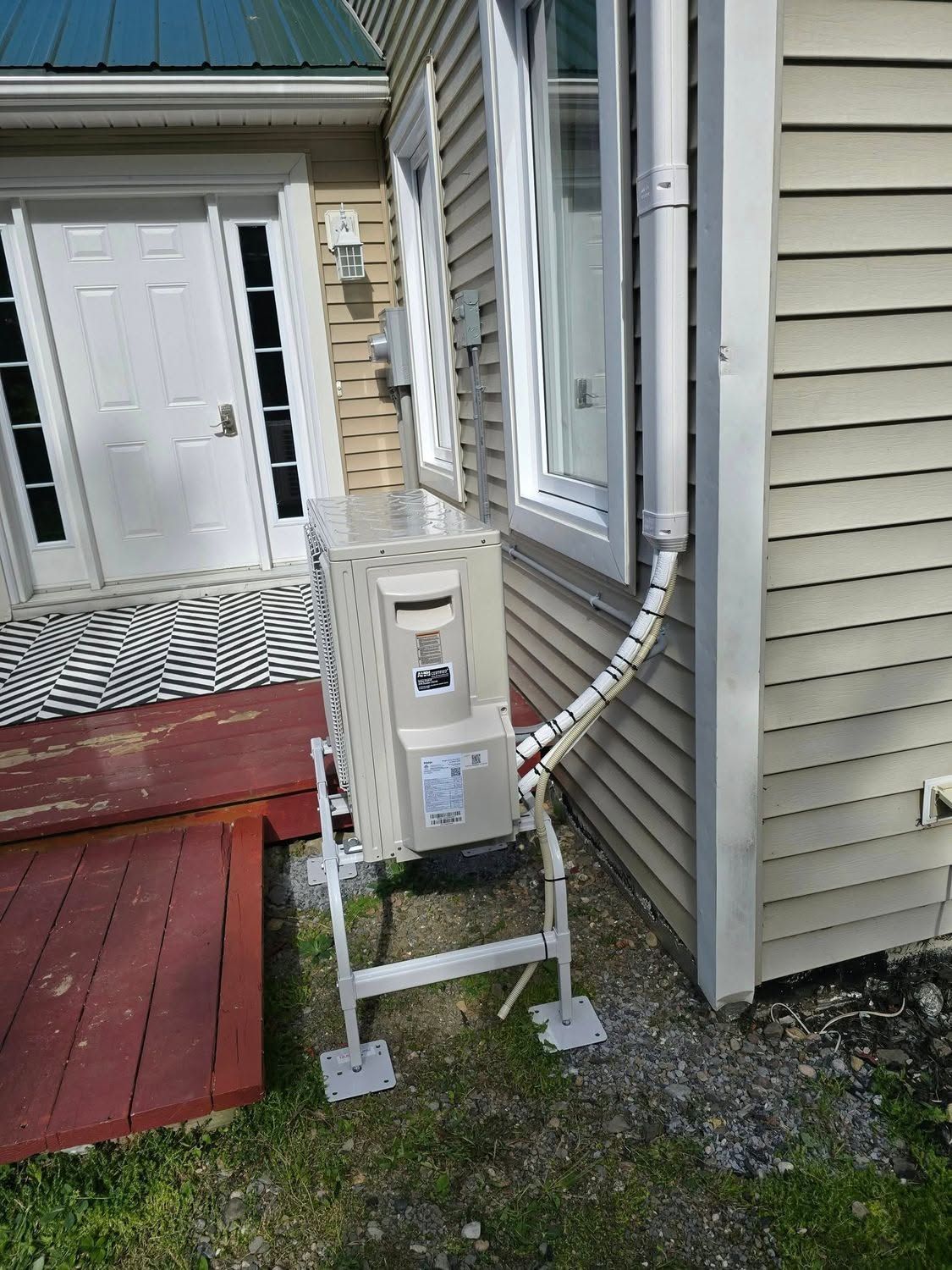 Portable HVAC unit outside house beside porch and white door, with duct hose attached.