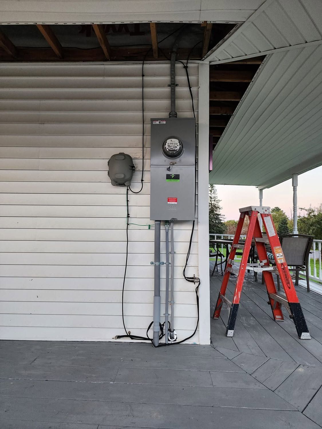 Exterior wall with electrical meter and gray panel box beside a red ladder under a covered porch.