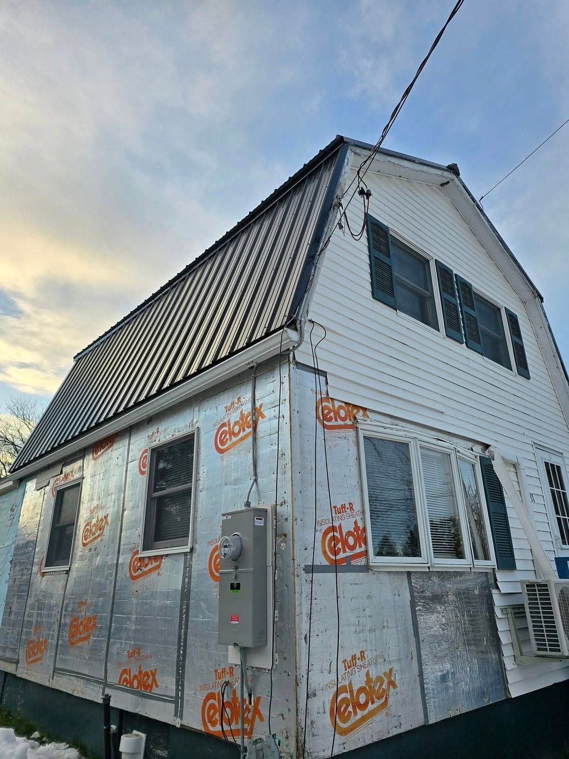Small house under renovation with silver house wrap, white siding, and dark roof at dusk