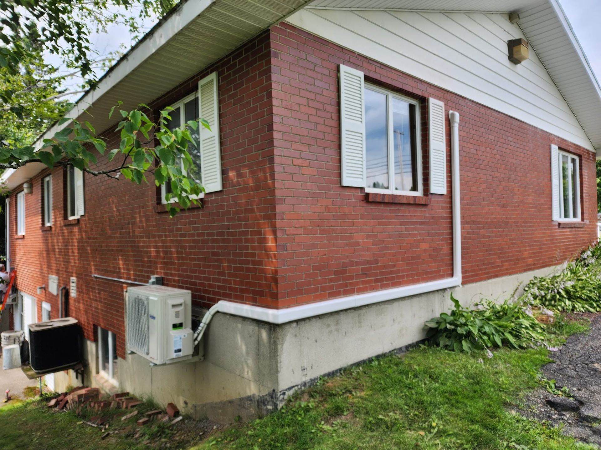 Red brick house exterior with white trim, basement windows, and air conditioning units beside a grassy yard.