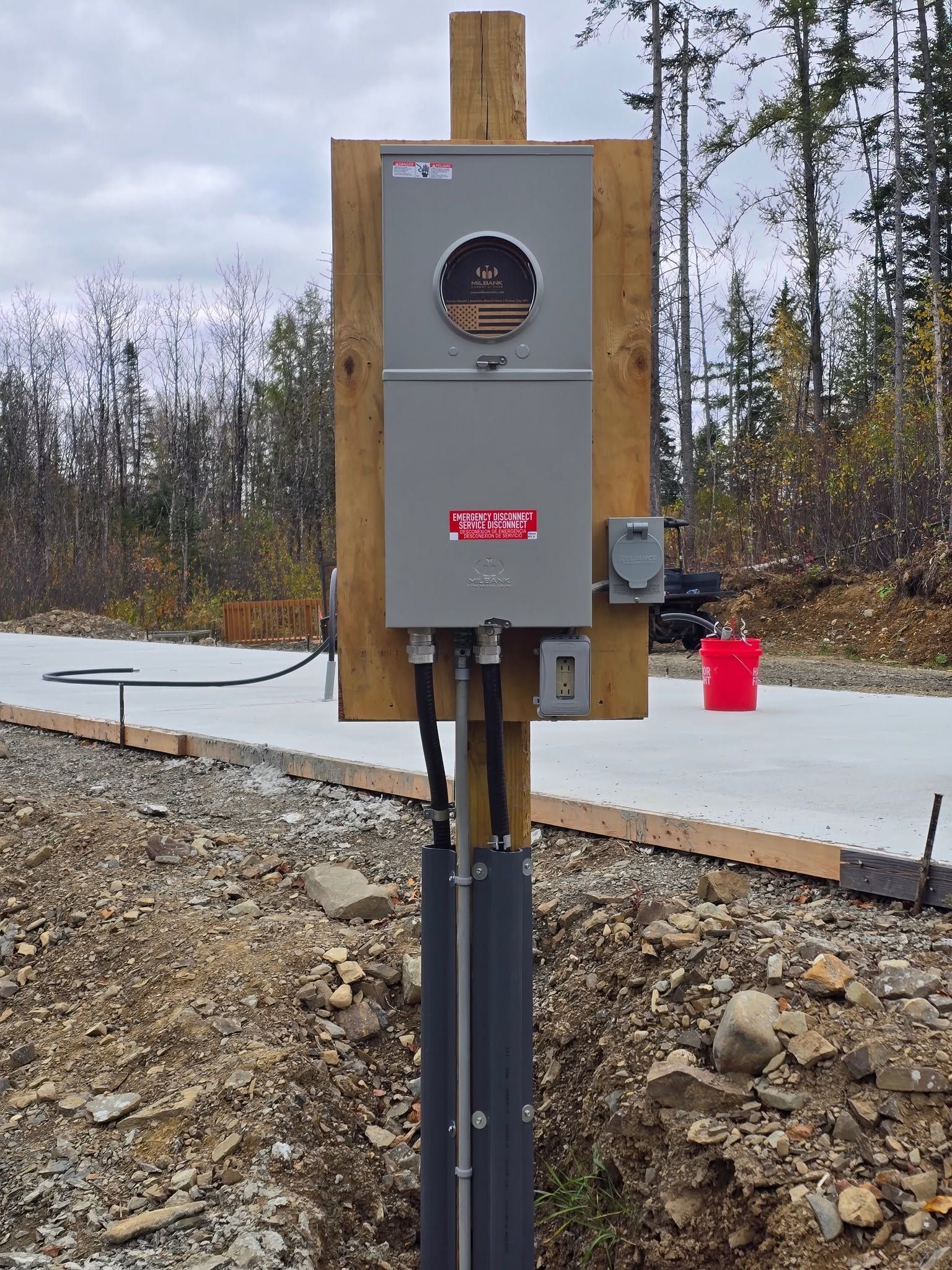 Gray electrical meter box mounted on a wooden post at a construction site, with pavement and trees behind it