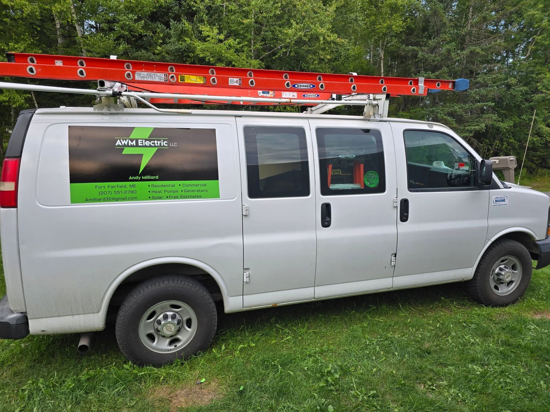 White utility van with a red ladder on top, parked on grass beside trees and a road.