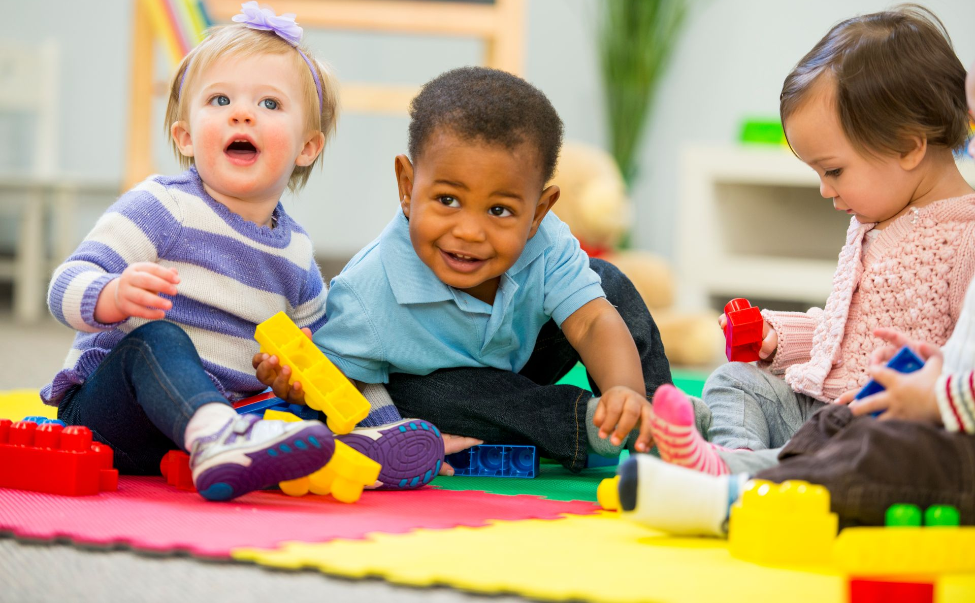 Three toddlers play with colorful toys on a foam mat, smiling in a bright playroom.
