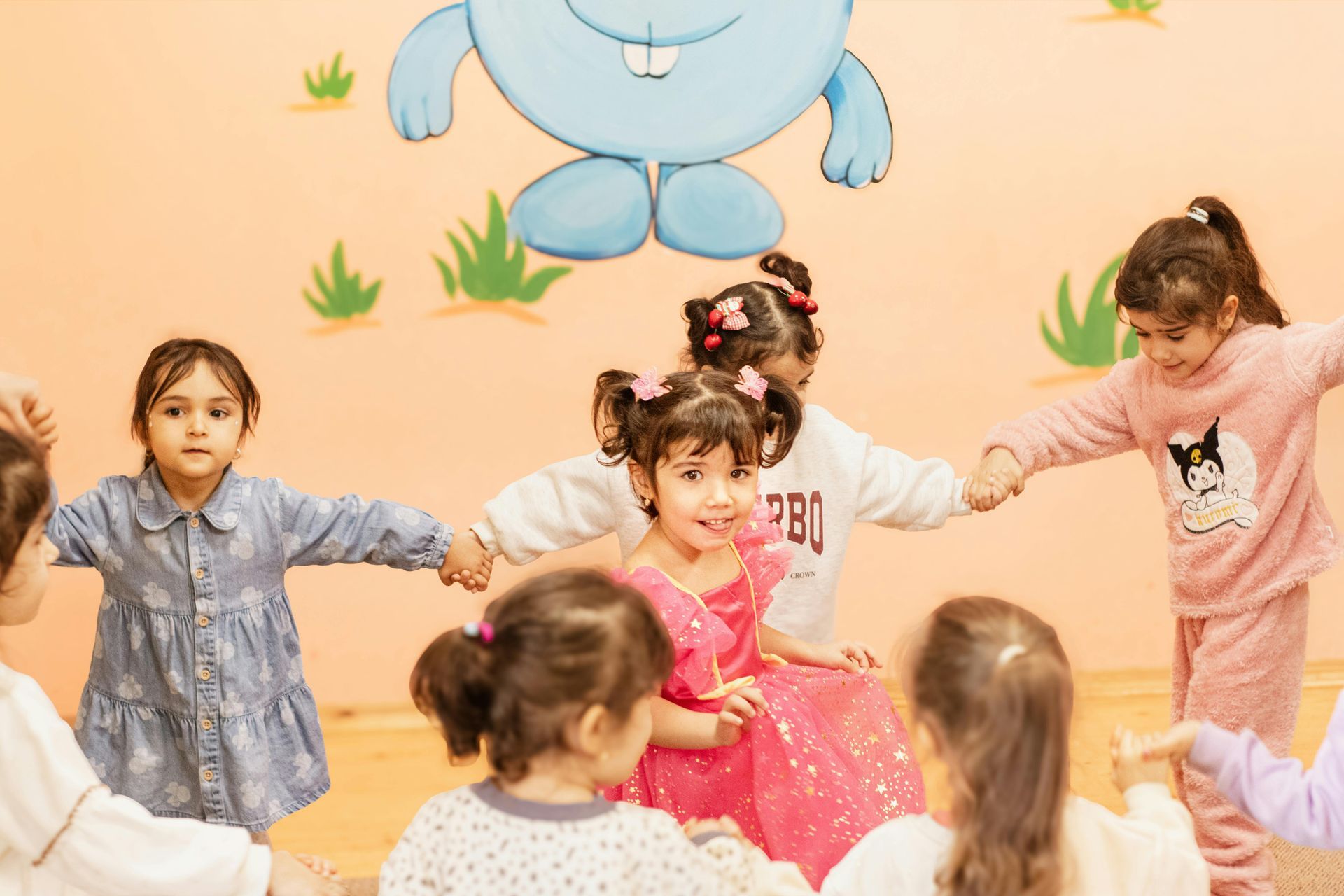 Children holding hands and dancing in a colorful classroom with a cartoon bear mural on the wall