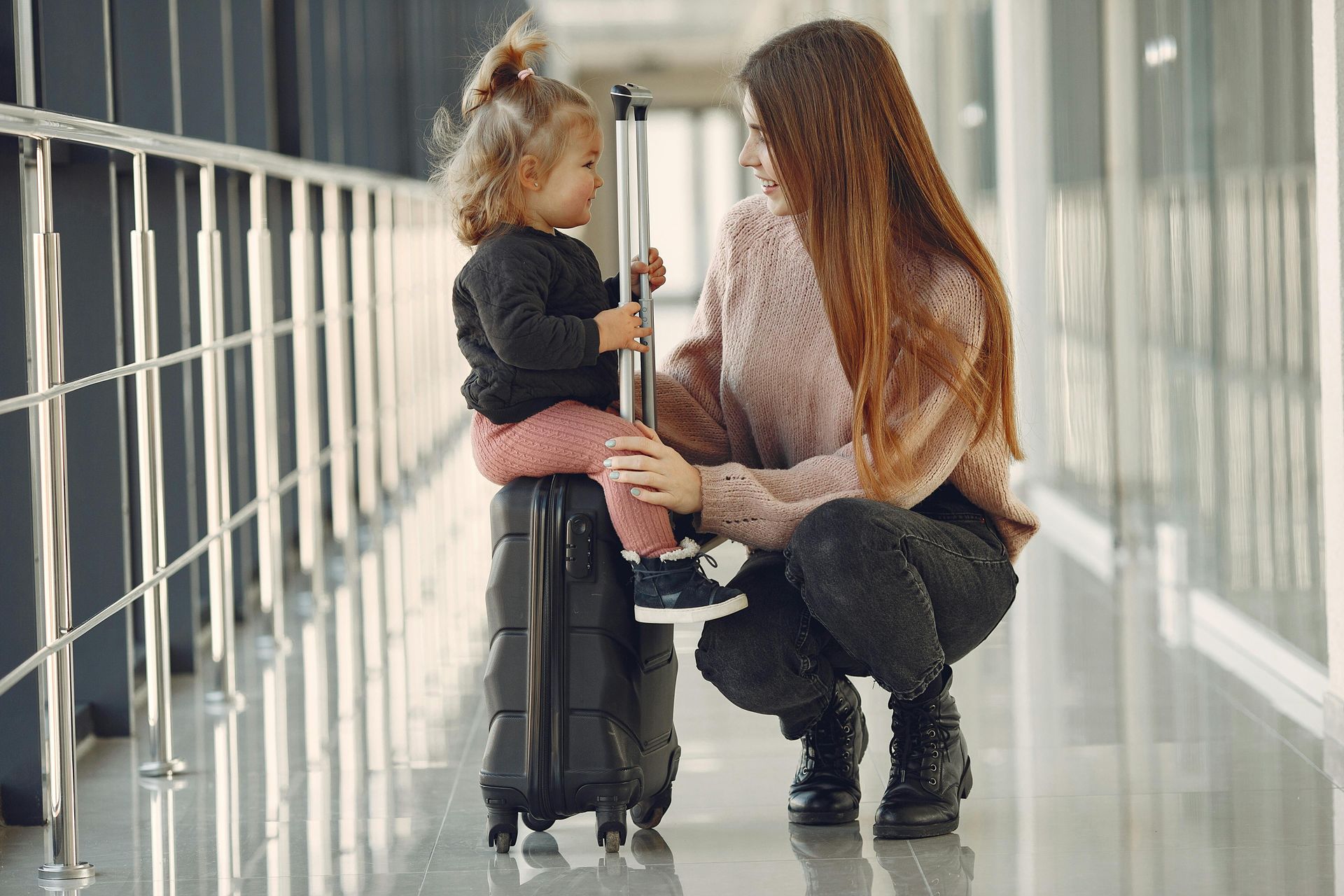 Woman kneeling to speak with a child in a bright hallway, holding the child’s hands.