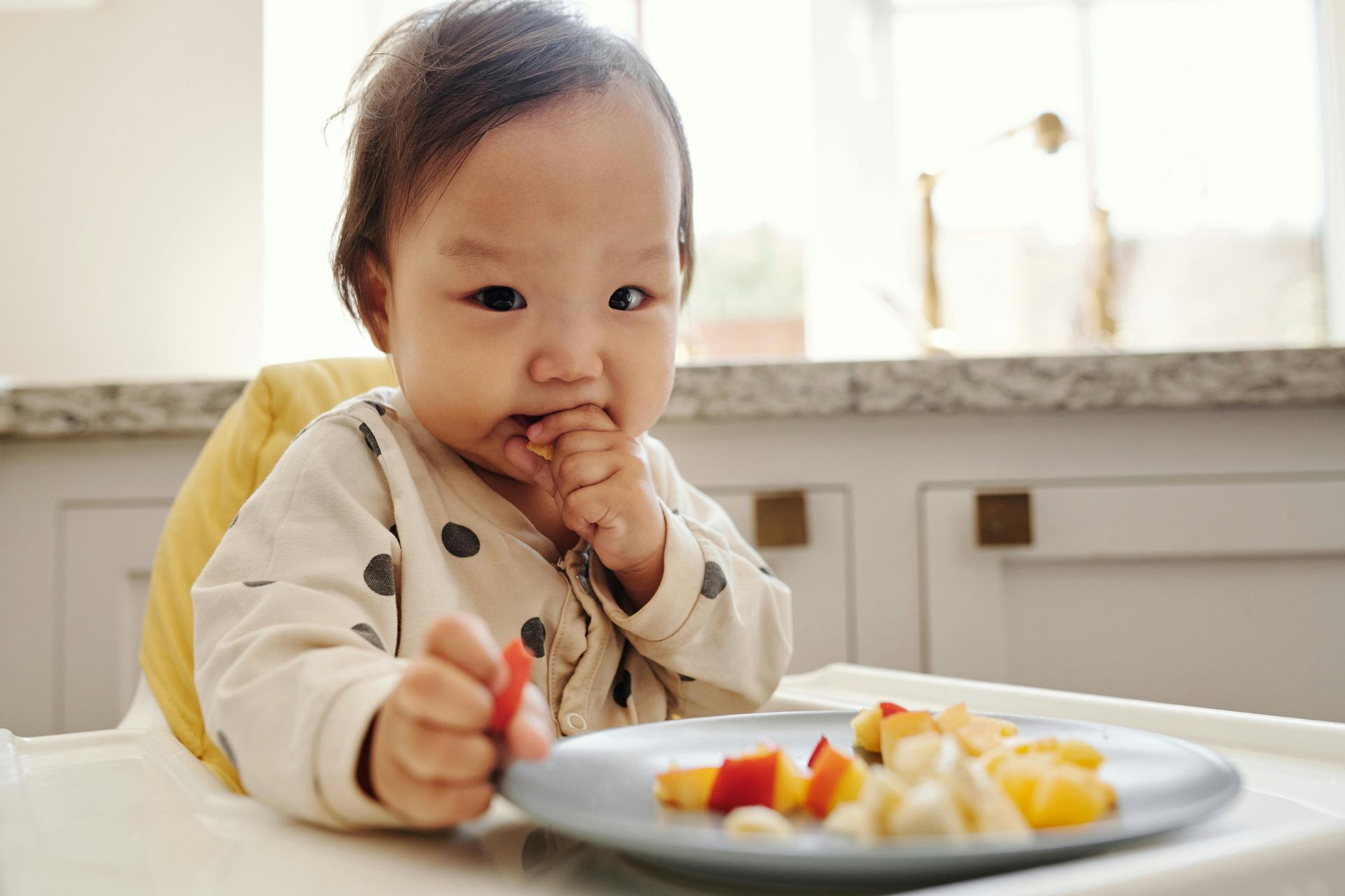 Child eating at a table, reaching toward a plate of fruit in a bright kitchen