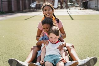Three children sitting on grass, smiling and stacked in a playful pose outdoors.