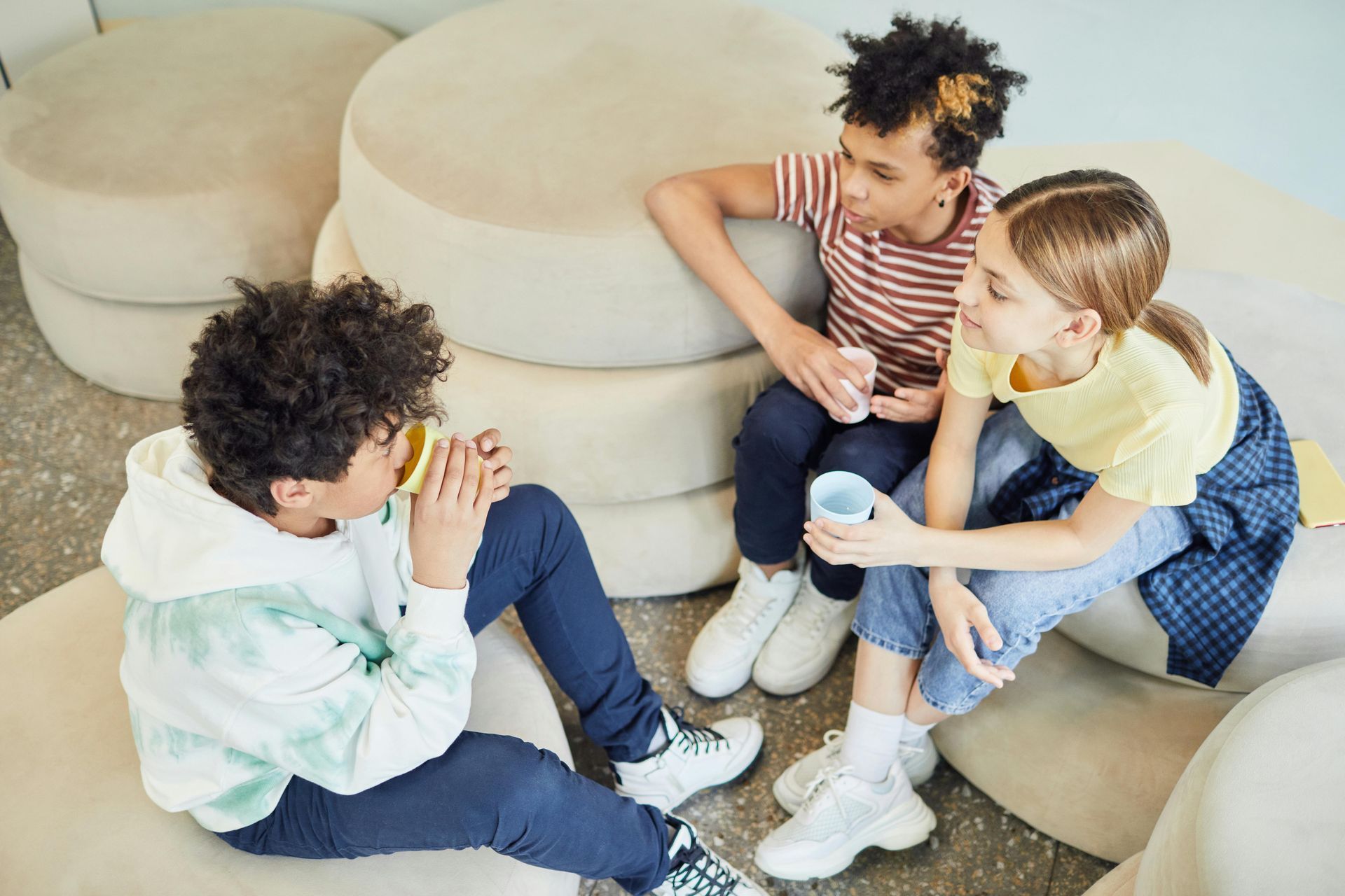 Three children sit together on round beige stools, sharing a tablet and chatting in a bright room.