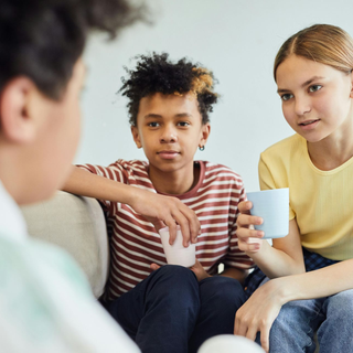 Three people sit together on a couch, one holding a white cup, in a casual conversation.