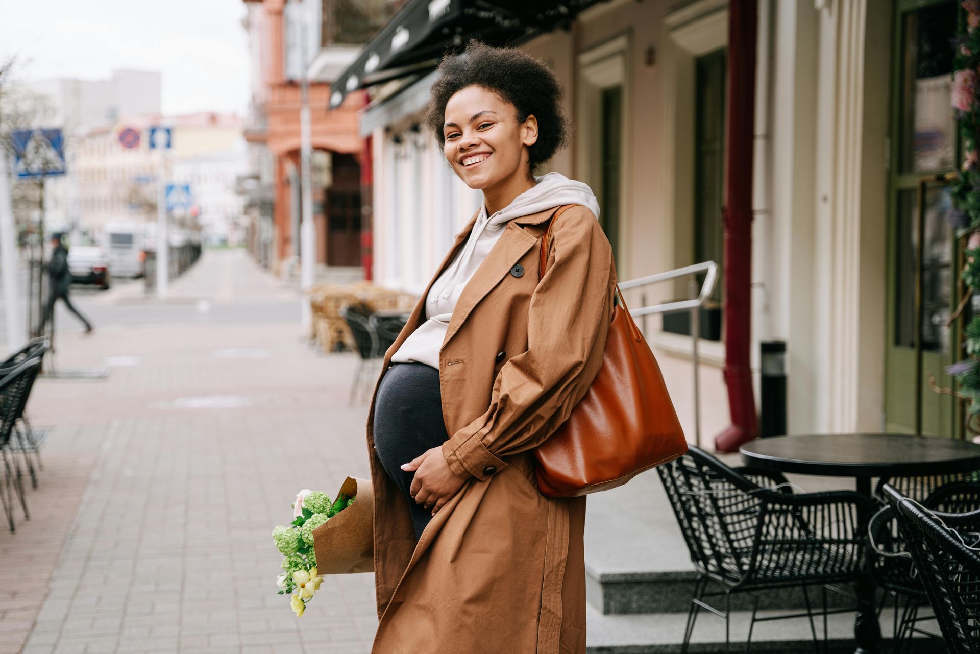 Smiling pregnant woman in a tan coat standing on a city sidewalk with a brown bag.