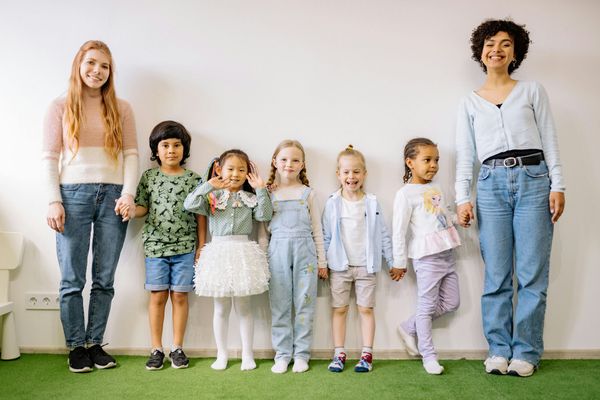 Six children standing in a row in a bright room on green carpet, holding hands and smiling