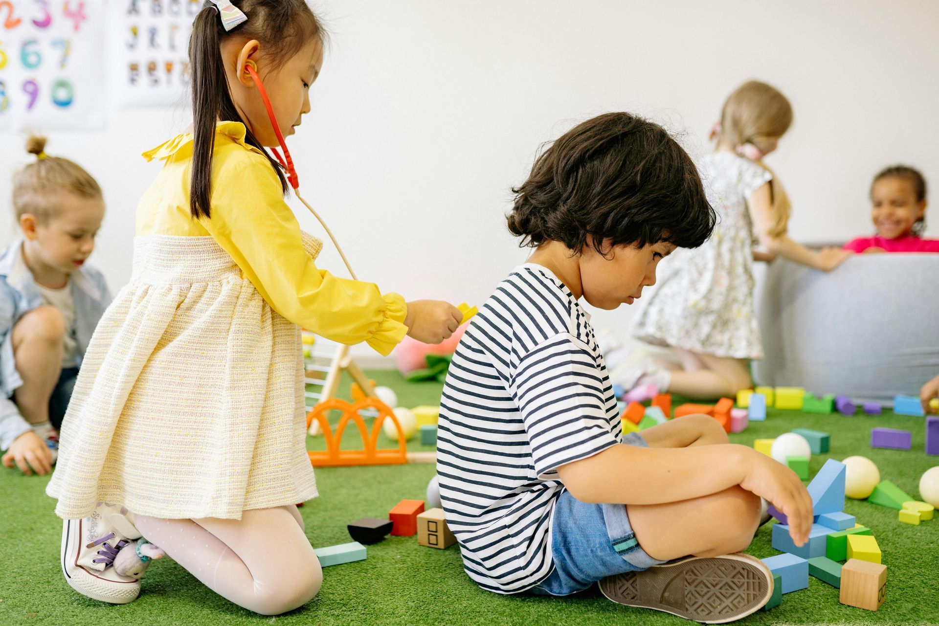 Children playing with colorful blocks on a classroom floor, with a teacher in the background