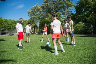 Children standing in a circle on a grassy field, playing soccer on a sunny day.