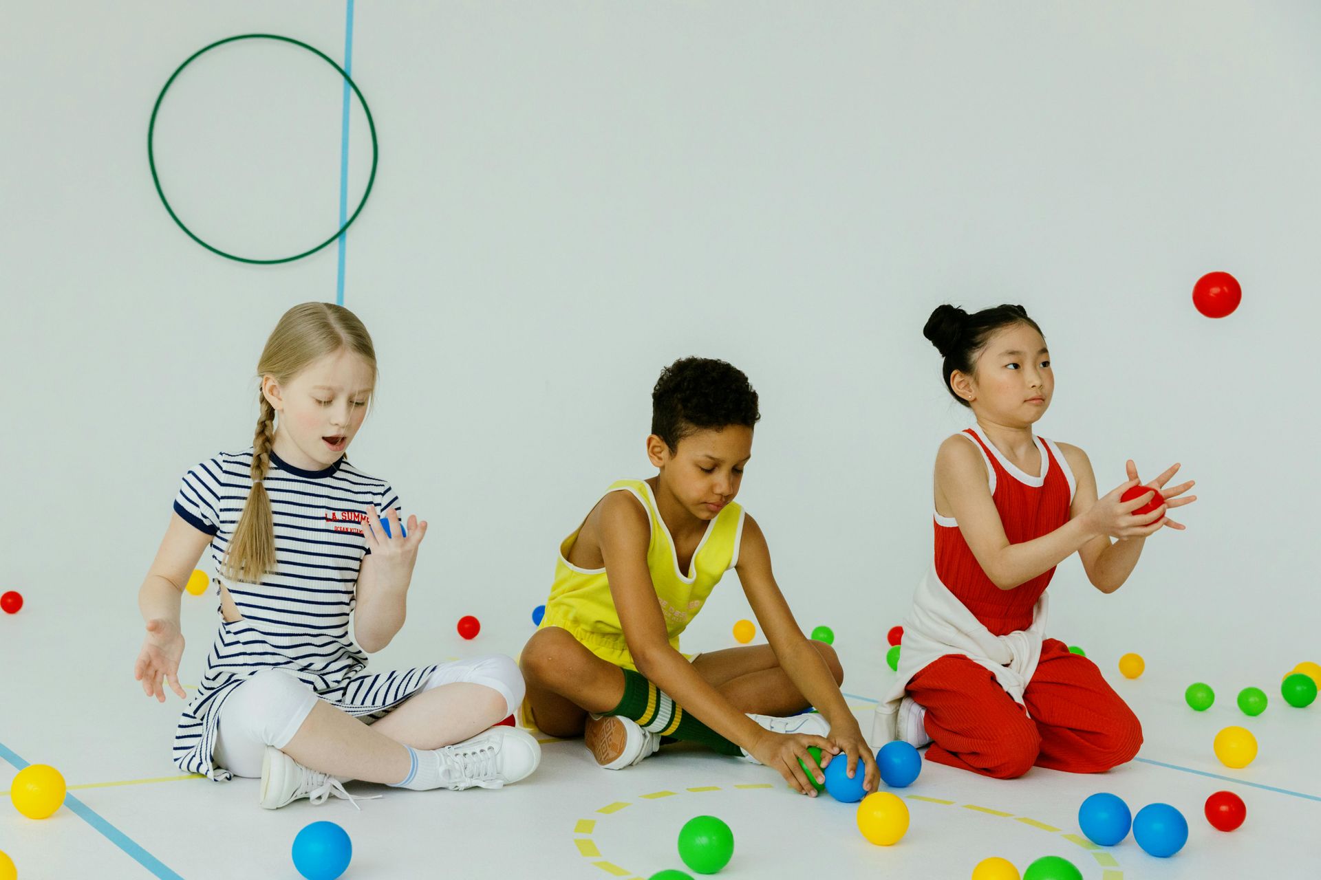 Three children sitting on the floor with colorful balls, playing in a bright room.