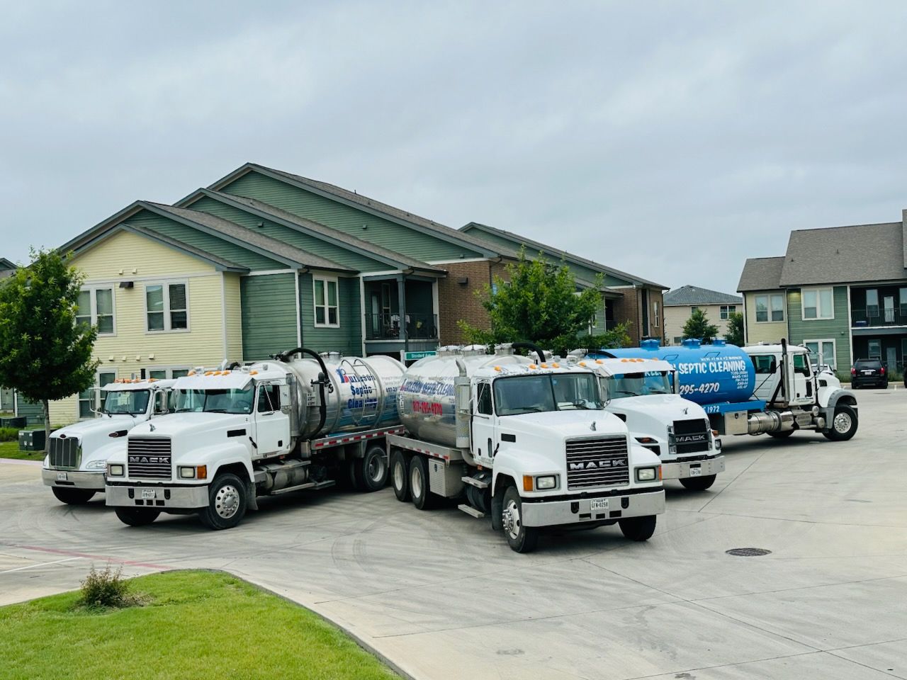 White tanker trucks parked in a residential street beside houses under a cloudy sky