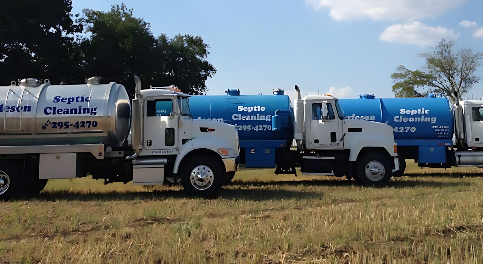 Two white and blue septic cleaning trucks parked in a grassy field under a clear sky