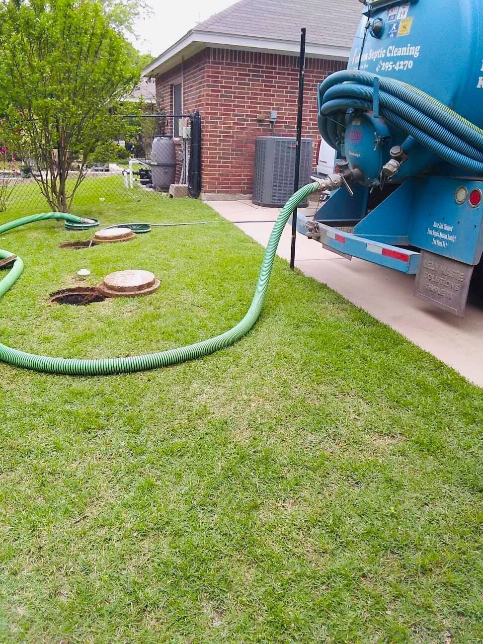 Blue sewer-cleaning truck hoses into a green lawn beside a brick house