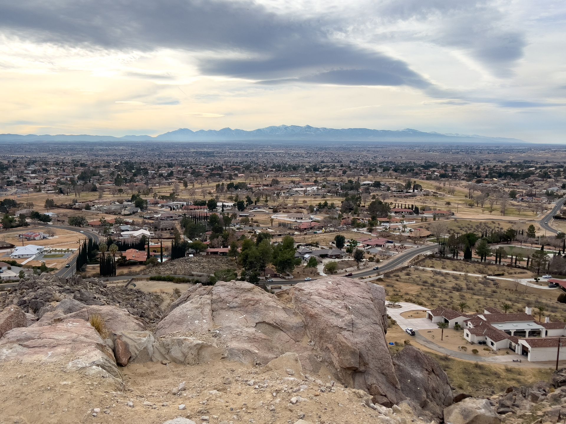 View of a city and distant mountains from a rocky, elevated location under a cloudy sky.