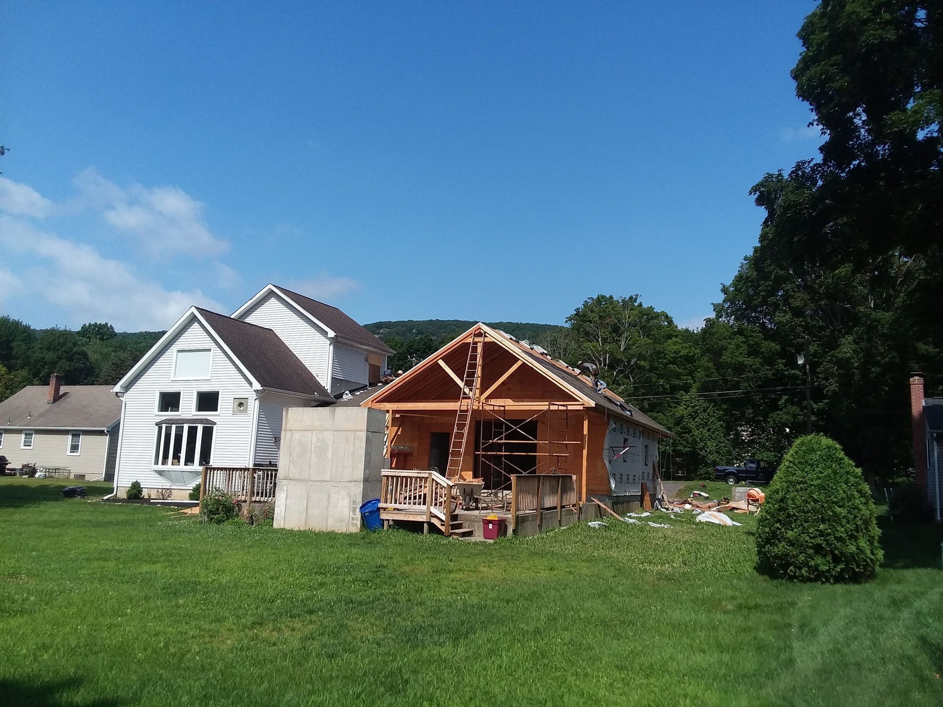 House under construction next to a white house, both on a grassy lawn with trees and a blue sky.
