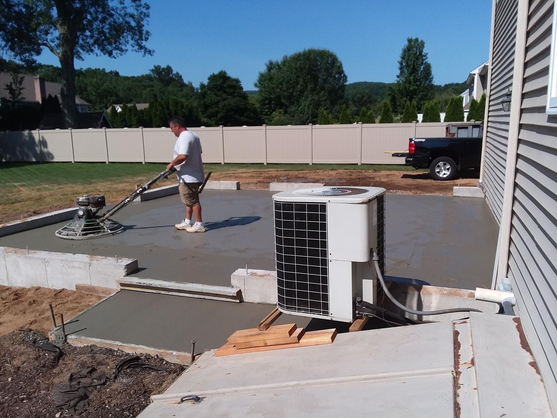 Man smoothing wet concrete with a power trowel on a patio next to a house with an air conditioner.