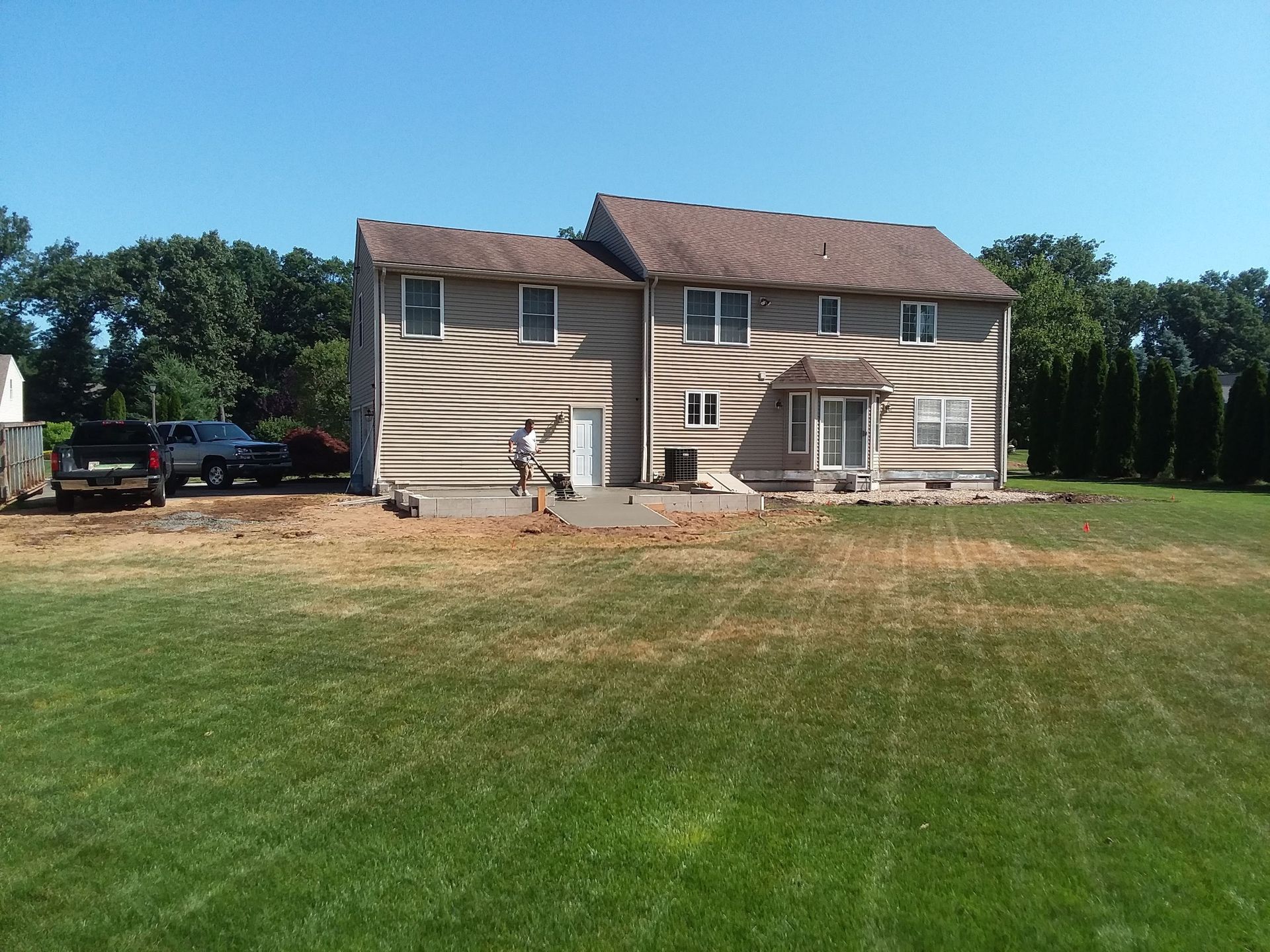Back of a two-story house with siding, surrounded by grass and trees; person on a patio near a door.