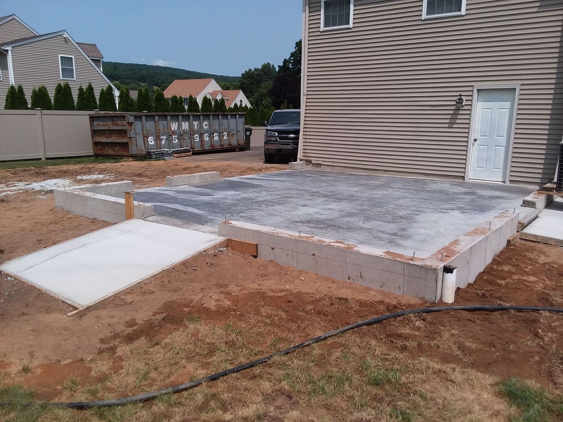 Concrete patio under construction next to a house with a door, driveway ramp and dumpster.