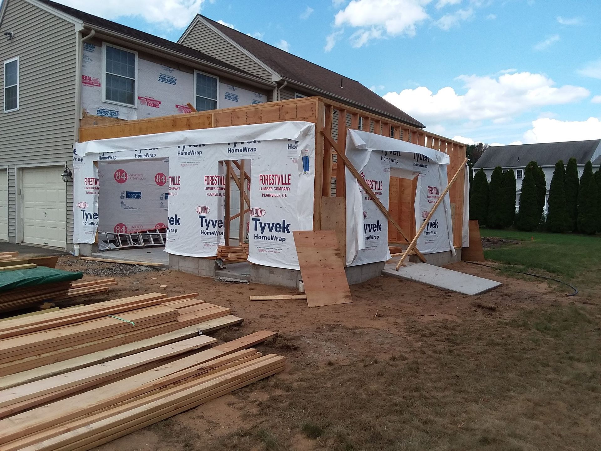 Construction of a home addition with exposed wood framing, covered in building wrap, set on a concrete foundation, outdoors.