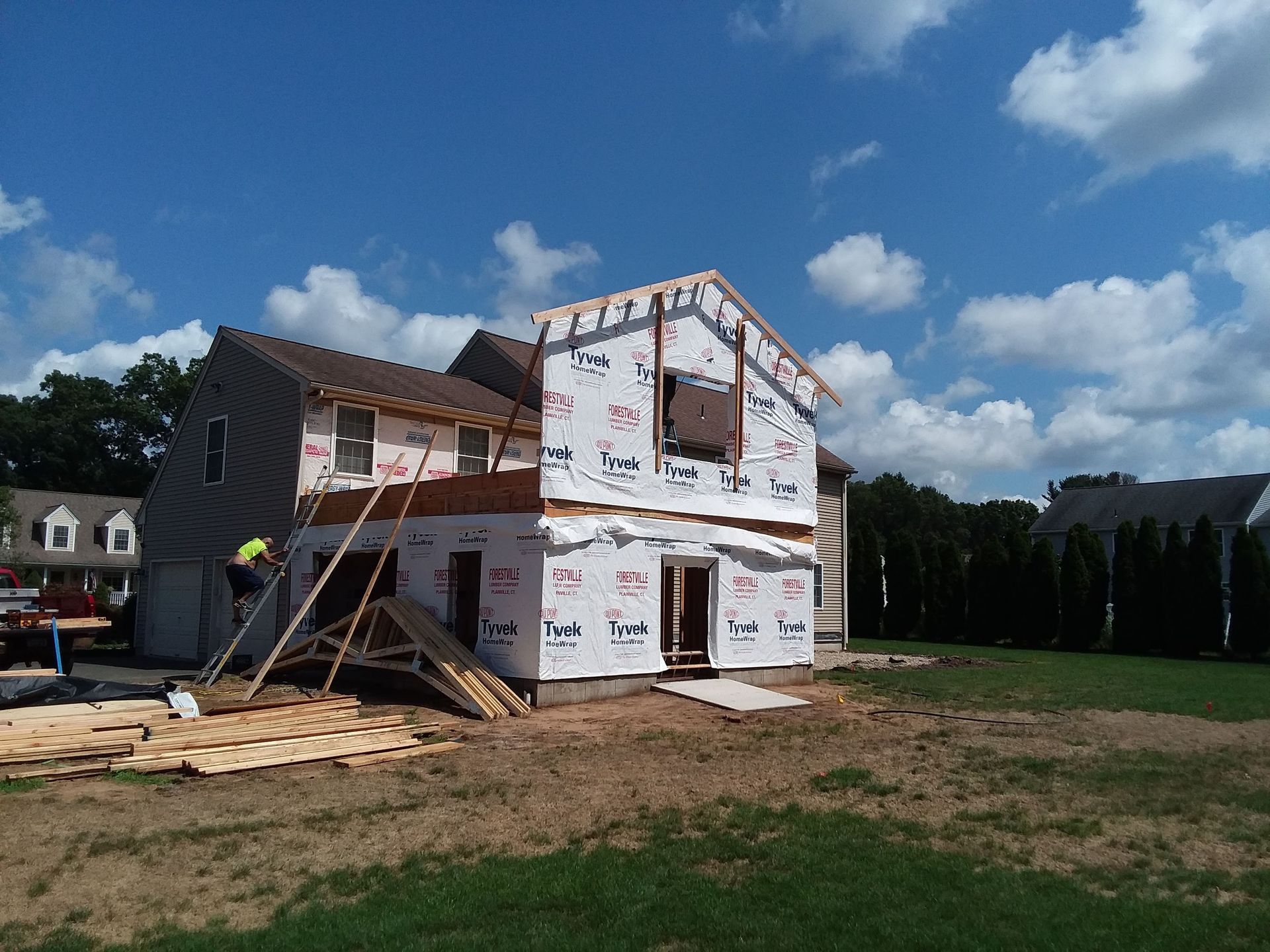 Two-story house under construction with a worker on a wooden frame; clear blue sky with some clouds.