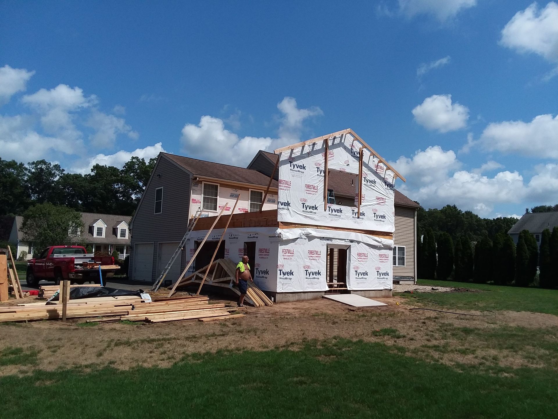 House under construction with worker, blue sky, and surrounding trees.
