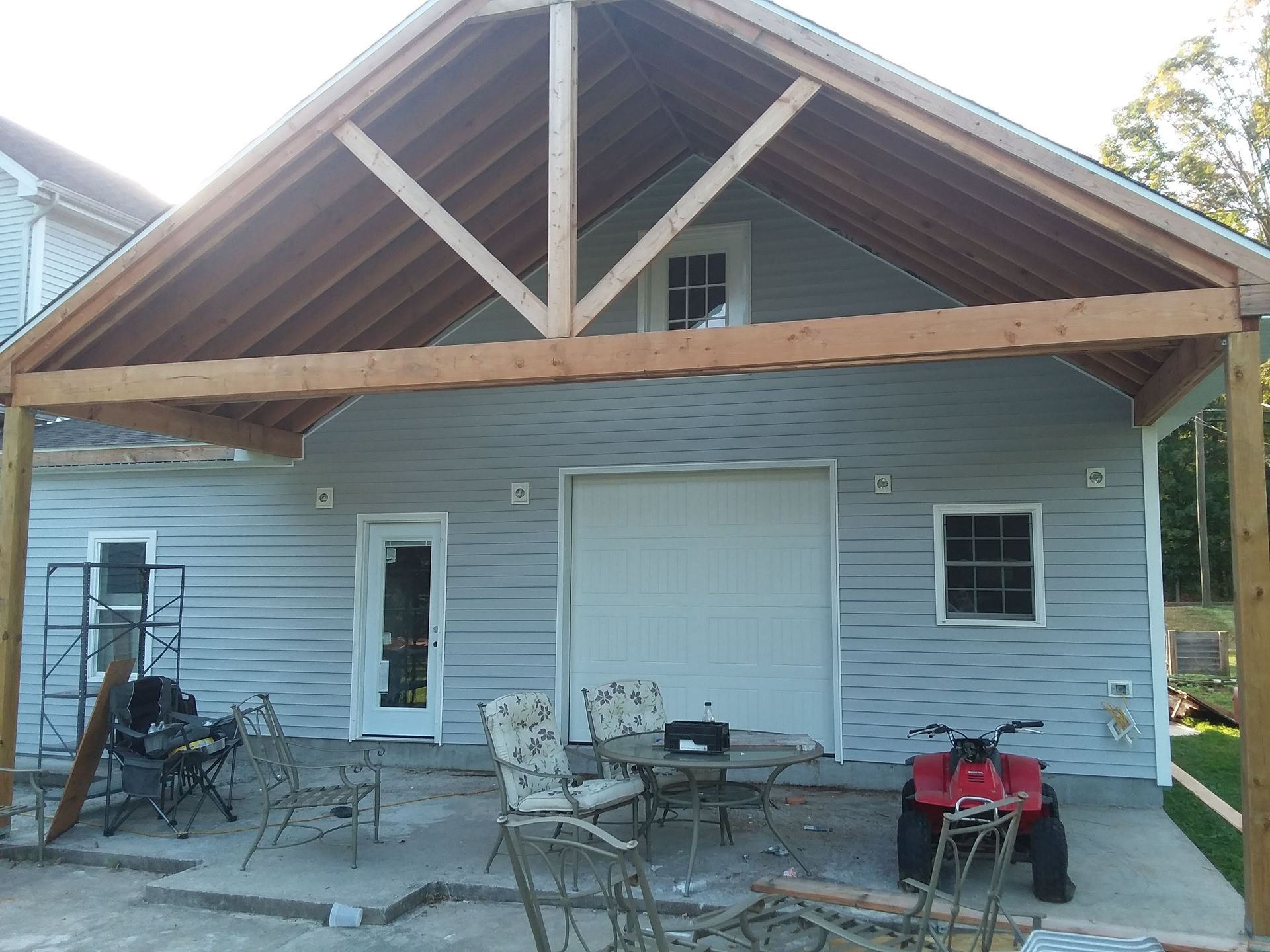 Attached wood-framed patio cover addition on a light blue-sided building with a garage door and small windows.