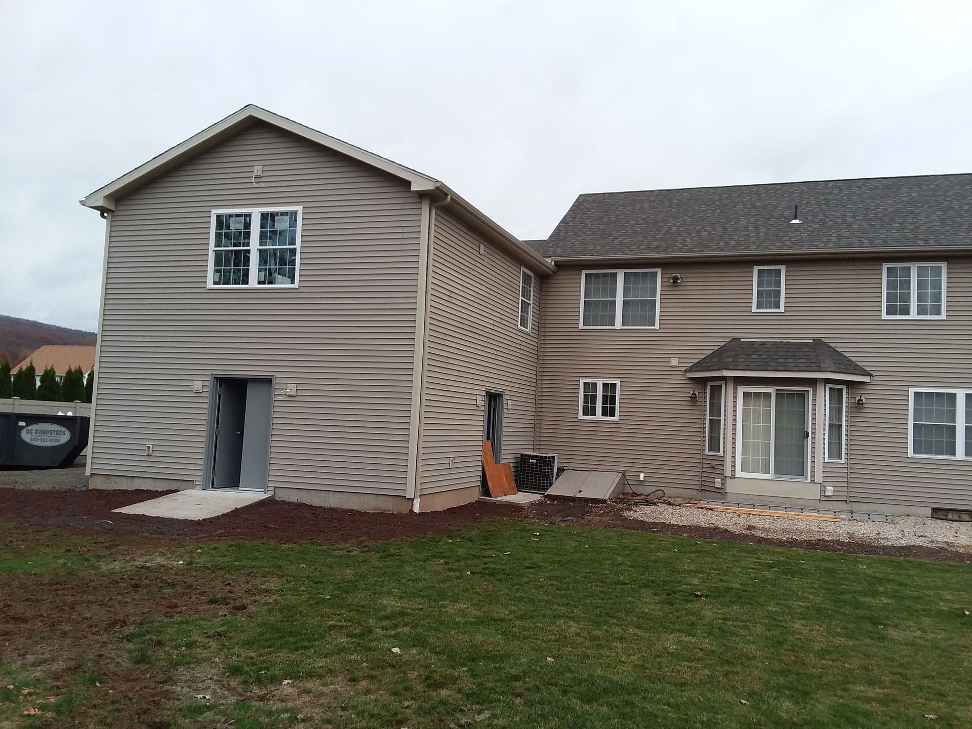 Backyard view of a house with a new addition. Beige siding, windows, and grass. Overcast sky.