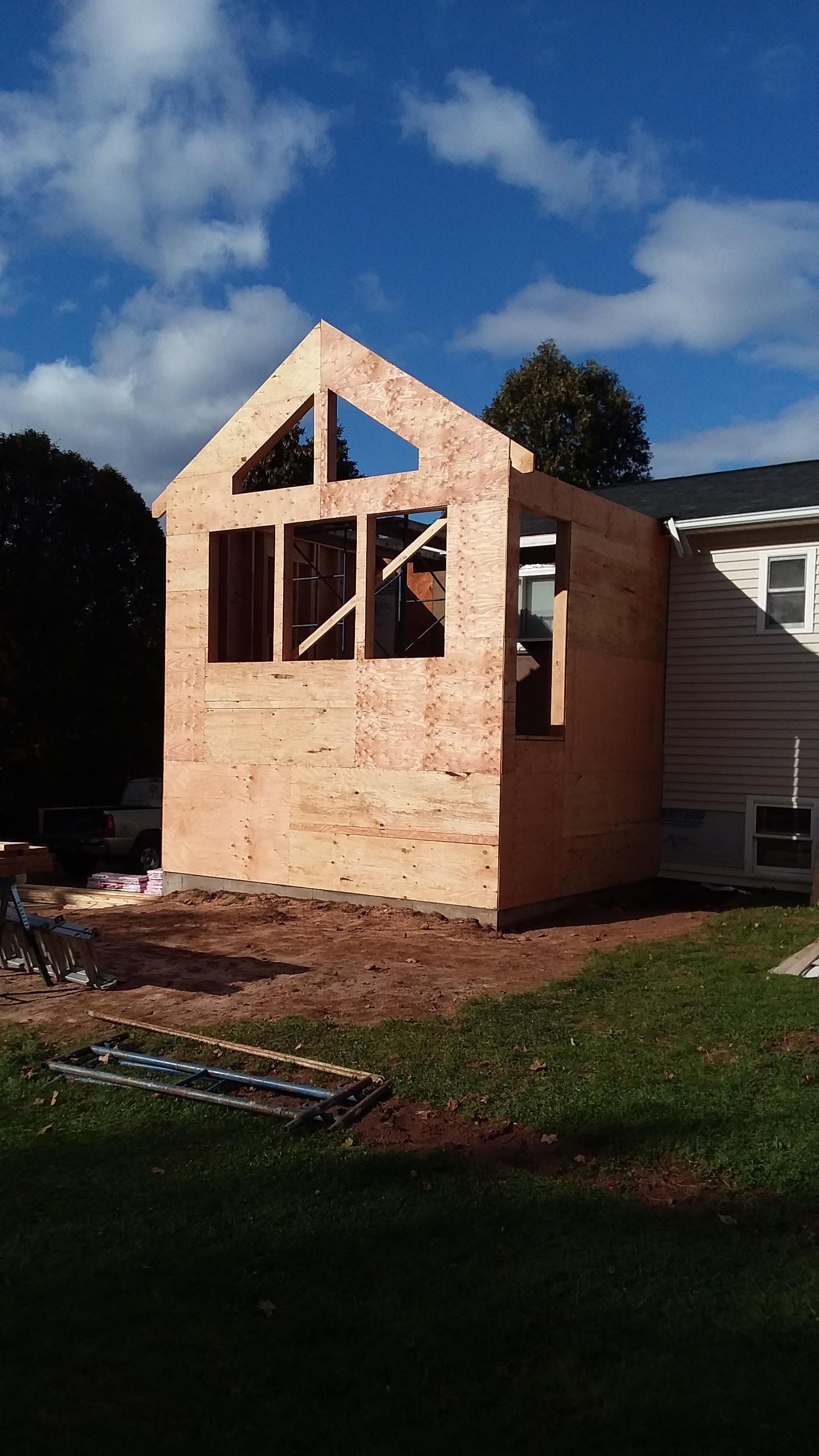Wooden frame of a house addition under construction, with a triangular gable roof, next to an existing house, on a sunny day.