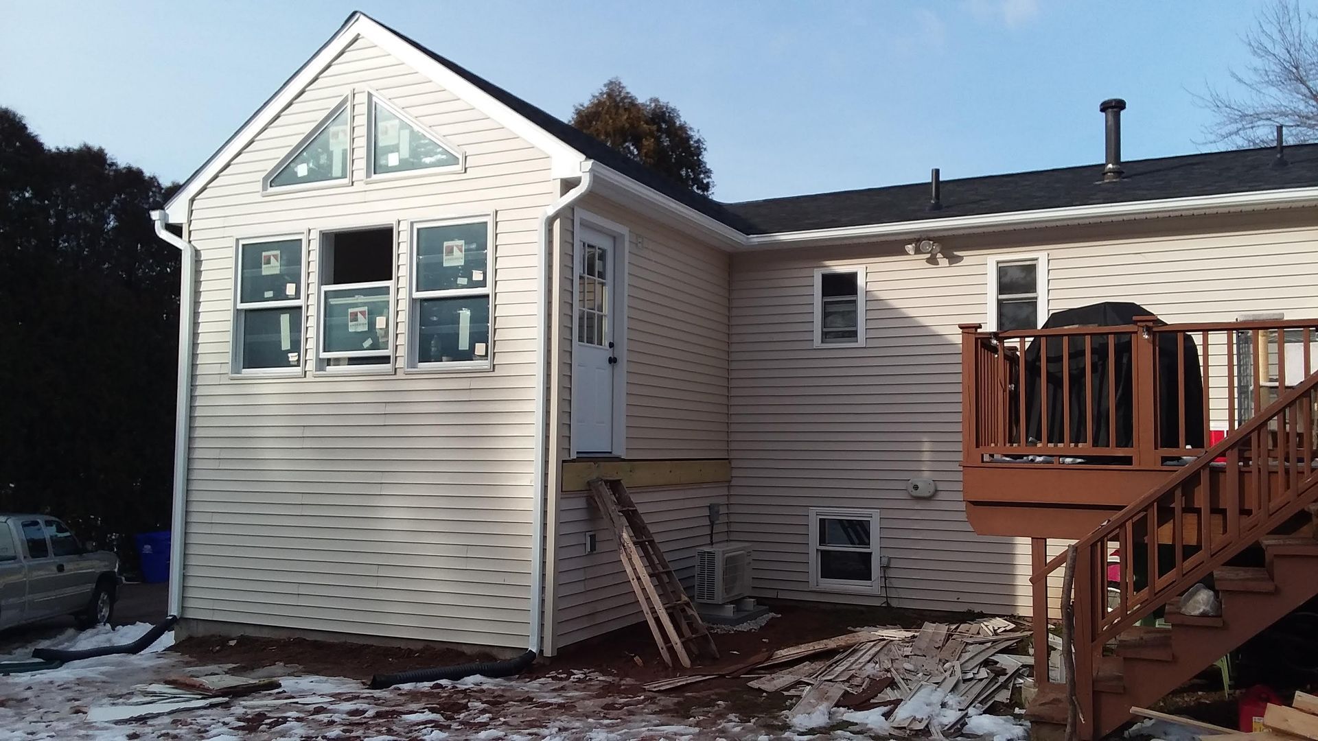 Exterior view of a house addition with new siding, windows, and deck under construction; snow on the ground.
