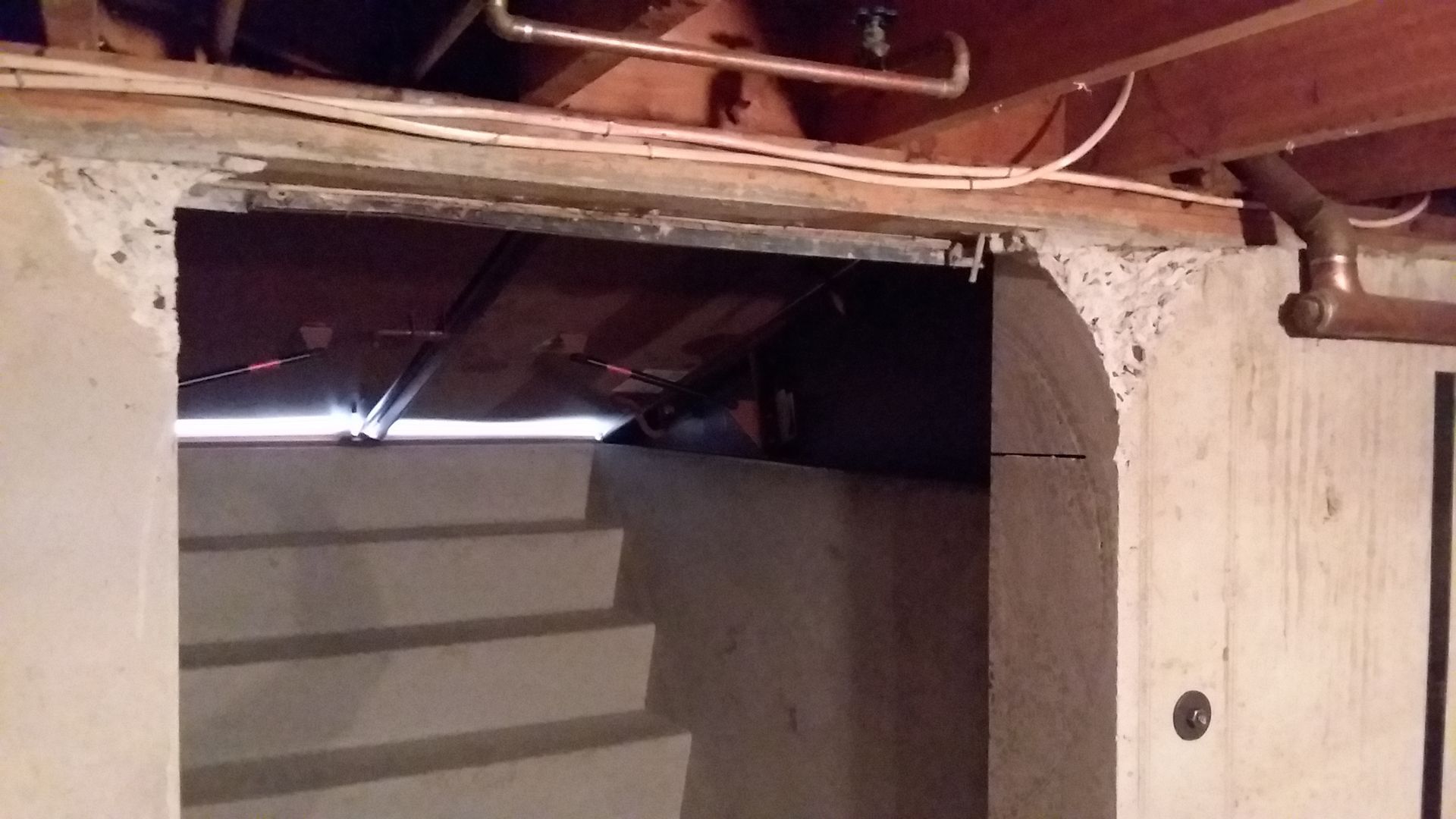 Concrete basement steps and doorway under wooden beams with exposed pipes.