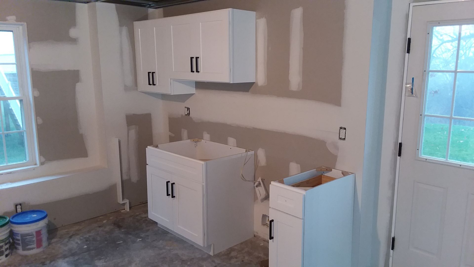 White kitchen cabinets installed in a room with unfinished walls; a window and door are visible.
