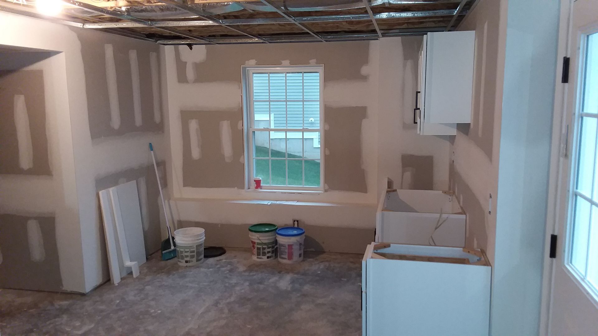 Kitchen under construction, with drywall, cabinets, and a window; ceiling exposed.